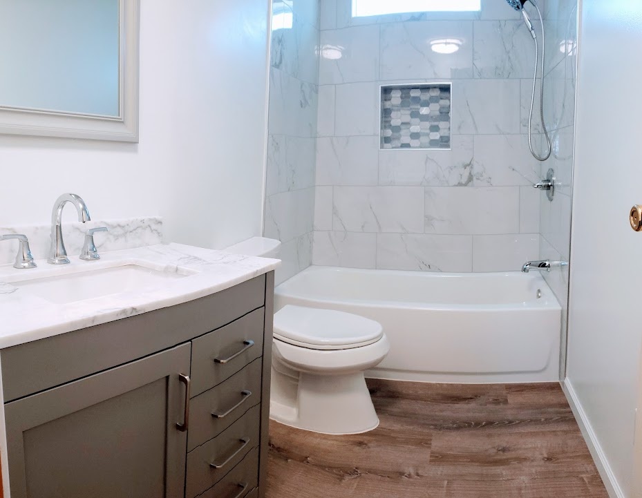 Finished bathroom with gray vanity, marble-look tile surround, inset shower niche, and wood-look flooring.